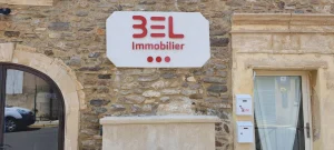 Panneau blanc BEL Immobilier aux lettres rouges sur une façade en pierre