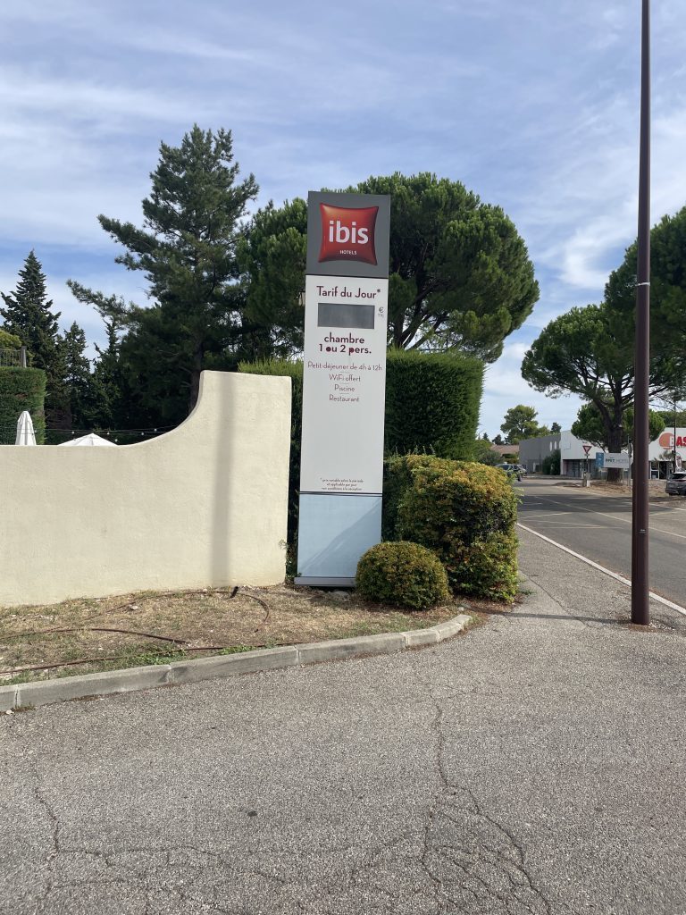 Totem d’entrée Ibis près du trottoir avec indication tarifaire et végétation en arrière-plan, pose totem Avignon