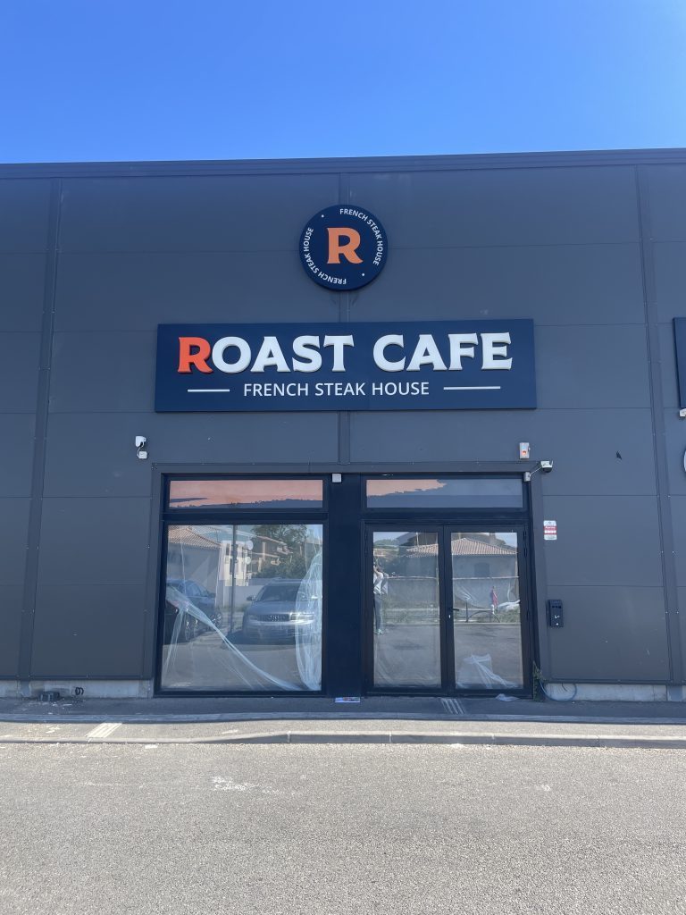 Façade grise montrant l’enseigne rectangulaire Roast Cafe orange et blanche et le logo rond au-dessus de la porte vitrée